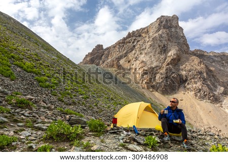 Traveling man eating meal Hiker sitting aside yellow camping tent and having lunch stove and cooking gear mountain landscape on background