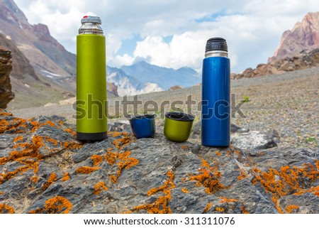 Two travel thermoses on stone.\
Green and Blue Thermos Flask with Opened Cups Located on Stone Mountain Landscape Background