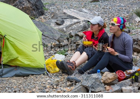 Outdoor Meal.\
Two Female Hikers Sitting along Camping Tent and Cooking Eating Smiling Talking