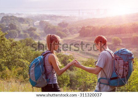 Young People Traveling.\
Girlfriend and Boyfriend Teenagers Couple with Backpacks on Green Forest Background Holding Hands Talking Smiling rising Sun on Background