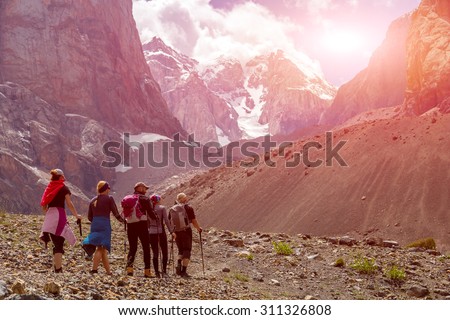 Group of Hikers Walking into Wilderness.\
Large Group of People Sport Clothing Going on Rocky Moraine Up towards Mountain Pass Peaks Sunlight Blue Sky Majestic Summits Background