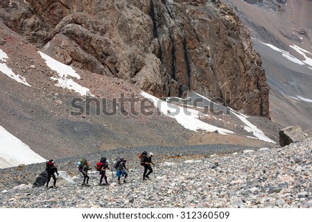 Extreme climbers scrambling up. Group people approach high altitude mountain climbing camp with heavy backpacks tons alpine gear walking on rocky path trail at peaks glaciers snow sunny day