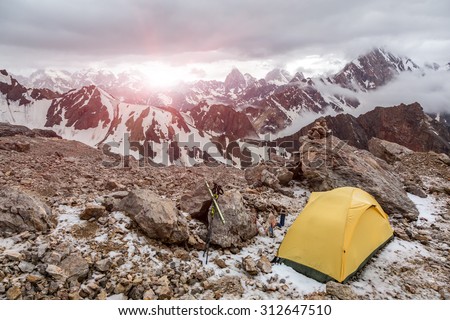Yellow tent on mountain landscape.\
Small Alpine camping Tent located on rocky terrain stone surface and high mountain hills and peaks on background evening sun light