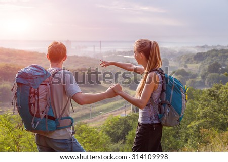 Romantic Journey Girlfriend and Boyfriend Teenagers Couple with Backpacks on Green Forest Background Pointing Hands Scenic View Smiling rising Sun on Background