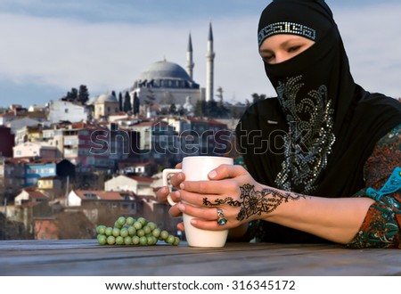Middle Eastern Woman at Cafe Terrace\
Turkish Lady Warming Hands Holding Tea Mug at Wooden Outdoor Cafe Table Traditional Clothing Istanbul Urban Landscape on Background focus on hand tattoo ornament