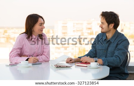 Man and Woman Casual Clothing Sitting at White Round Table with Coffee Mugs Laptop Notepads and Telephone Discussing Smiling Making Hand Notes Urban Landscape on Background