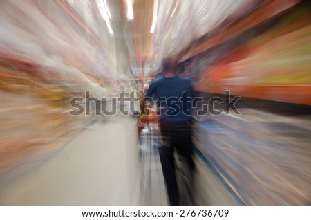 Male shopper pushing a shopping cart in a large grocery store. Image is blurred to imply motion, activity, and stress.
