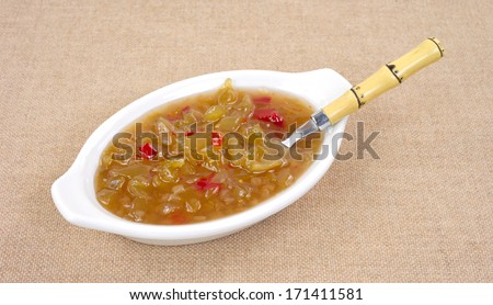 A small white dish with green tomato piccalilli and a spoon on a tan cloth background.