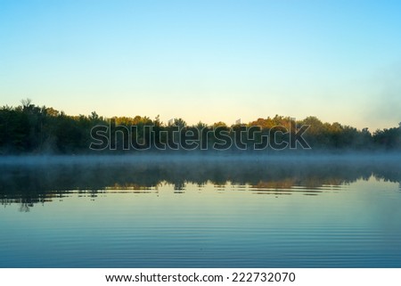 A calm early morning mist hanging above the water with the sun just touching the tops of the forest and trees at Toddy Pond, Maine.