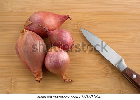 A group of shallots ready to be sliced on a wood cutting board with a knife to the side.