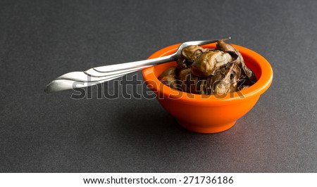 A small orange bowl filled with small whole smoked oysters with a fork on a black table top.