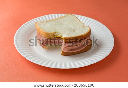 A bitten bologna sandwich with mustard and white bread on a paper plate atop an orange table top.