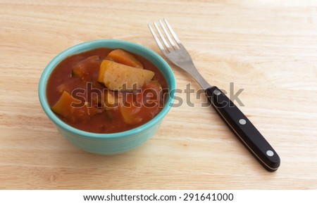 A small bowl filled with cooked zucchini in a tomato sauce on a wood table top with a fork to the side illuminated by window light.