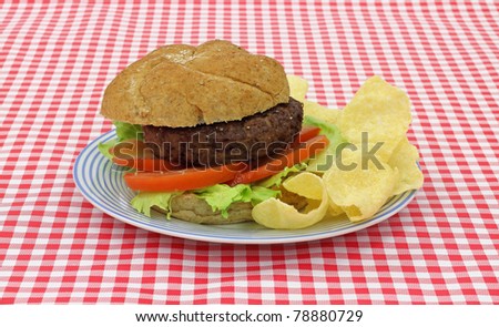 A freshly cooked low fat hamburger on a whole wheat bun with lettuce and tomatoes on a blue stripped plate with potato chips on red white checkerboard cloth.
