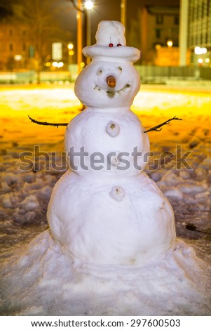 Happy snowman by night in the garden against buildings of the city.