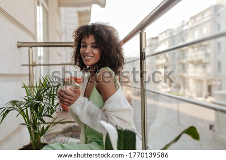 Happy brunette woman drinks coffee on balcony. Charming lady in light green dress smiles sincerely on terrace.