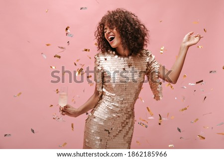Happy woman with wavy dark hair style in shiny trendy clothes rejoicing, holding glass with champagne and posing with confetti on pink backdrop..