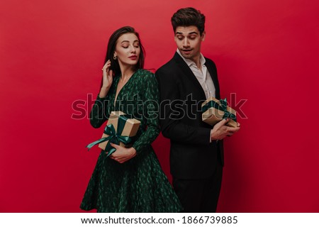 Lovely pair of people wearing dark elegant clothes, standing back to each other and holding gift boxes against red plain background  ストックフォト © 