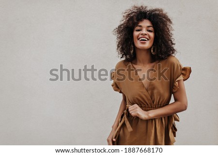 Brown-eyed woman in brown dress smiles on grey background. Stylish brunette curly dark-skinned lady moves on isolated.