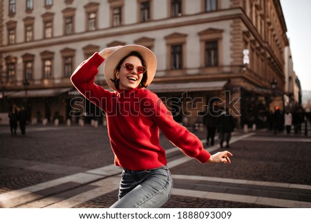 Cool girl in red sweater looks away. Attractive short-haired woman in beige hat and sunglasses walks outdoors.