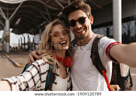 Young attractive blonde woman in plaid shirt and stylish brunette man in sunglasses smiles and takes selfie. Portrait of couple of travelers near airport.