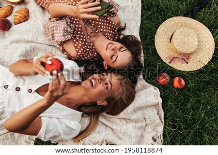 Charming attractive blonde and brunette girls in summer dresses lay on linen rug and have picnic. Cool woman in polka dot dress holds phone.