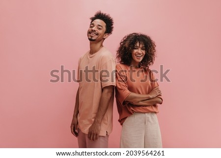 photo below belt of cheerful people of exotic appearance on isolated nacre pink wall. brunette with her arms crossed dressed in carrot-colored T-shirt tucked in jeans. guy in peach t-shirt.
