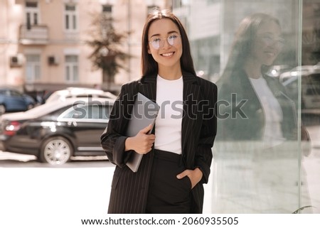 Brunette business young asian woman with laptop in her hands not in full growth smiling at camera. Lady with glasses stands outside office building during her lunch break. Сток-фото © 