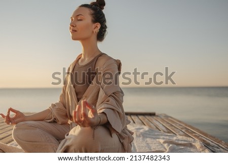 Beautiful modern young woman meditates with closed eyes, relaxes her body and clears her mind. Brown-haired woman with bun on her head is dressed light brown jacket and pants. Rest time concept. 商業照片 © 