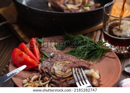 two fried marbled beef steaks with fried onions, tomatoes and dill on wooden background, rustic. Focus on piece of meat strung on fork, beer poured into glass, selective focus.Healthy food concept Imagine de stoc © 