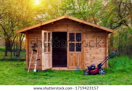 Hovel after work in evening, golden hour. Garden shed (front view) with hoe, string trimmer,  rake and grass-cutter. Gardening tools shed. Garden house on lawn in the sunset. Wooden tool-shed.  商業照片 © 
