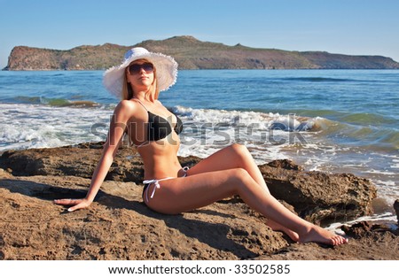 Young smiling blond caucasian white woman wearing black bikini, hat and sunglasses.Model lying on a beach, looking at viewer.