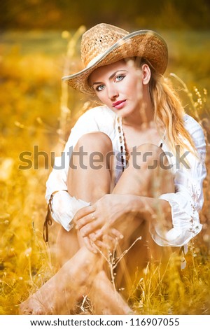fashion portrait woman with hat and white shirt sitting on a hay stack.very cute blond woman sitting down outdoor on the yellowgrass with a hat
