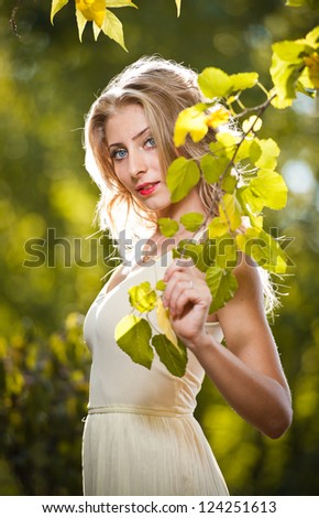 Young attractive woman in a romantic autumn scenery. Blonde woman posing in white dress in autumn scenery.Girl with leaves in hand and fall yellow maple garden background