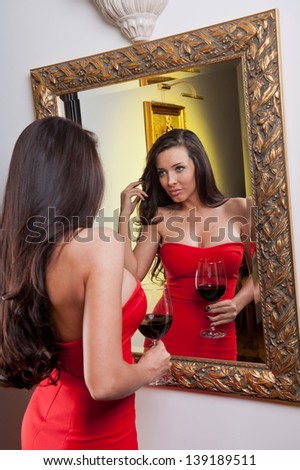 The beautiful girl in a long red dress posing in a vintage scene.Young beautiful woman wearing a red dress in the old hotel.Sensual elegant young woman in red dress looking into mirror