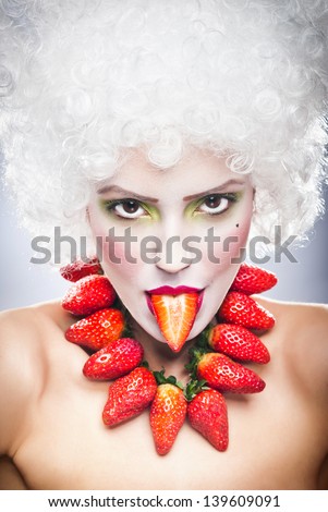 Creative makeup beauty shot of model with strawberries, artistic edit .Woman with strawberry necklace, wig and makeup professionally posing in studio.Beauty with strawberry.