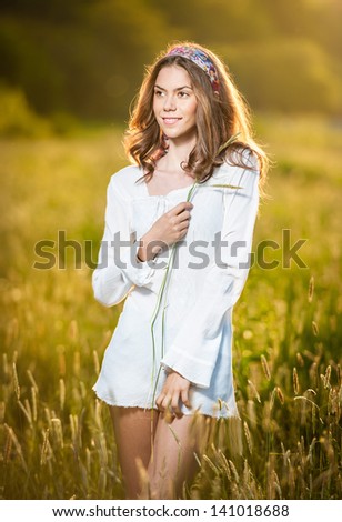 Girl with white shirt on the field at sunset.Young beautiful woman standing in wheat field .Vintage looking picture of a Young woman standing in a wheat field .Sun at early sunset in the background.