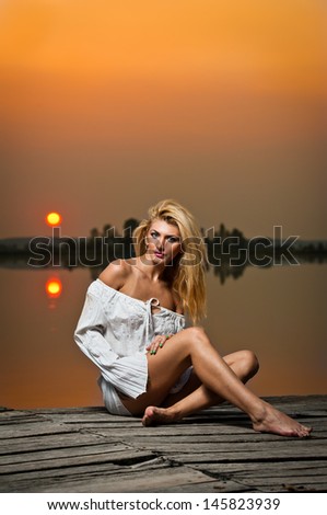 beautiful girl with a white shirt on the pier at sunset.Sexy woman with long legs sitting on a pier .Color image of a beauty girl sitting on a pier, overlooking a lake