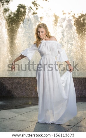 Attractive girl in whitelong dress sitting in front of a fountain in the summer hottest day. Girl with dress partly wet posing near a fountain. Blonde women near the fountain in a ballet position