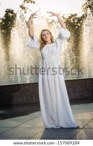 Attractive girl in white long dress sitting in front of a fountain in the summer hottest day. Girl with dress partly wet posing near a fountain. Blonde women near the fountain in a ballet position