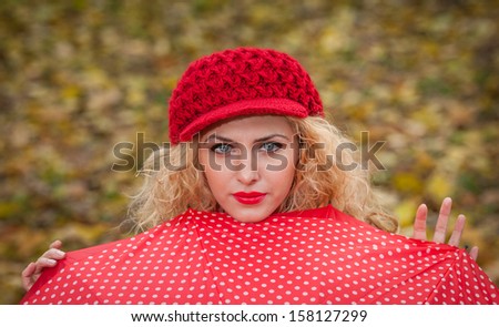 Attractive blonde girl with red cap looking over red umbrella outdoor shoot. Attractive young woman in a autumn fashion shoot.