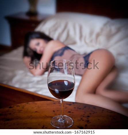 Mysterious lady laying in bed with a glass of red wine foreground. Sensual woman on bed and glass of wine. Beautiful girl in sensual lingerie posing provocatively indoor,  relaxation moments