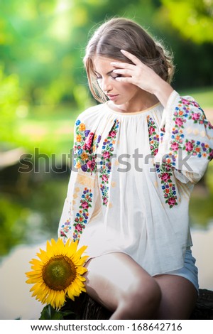 Young girl wearing Romanian traditional blouse holding a sunflower outdoor shot. Portrait of beautiful blonde girl with bright yellow flower. Beautiful woman looking at a flower harmony concept
