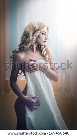 Young blonde woman wrapped in white towel posing relaxed. Beautiful young woman with a towel around her body after bath. Side view of long fair hair girl sitting on chair exposing her shoulder.