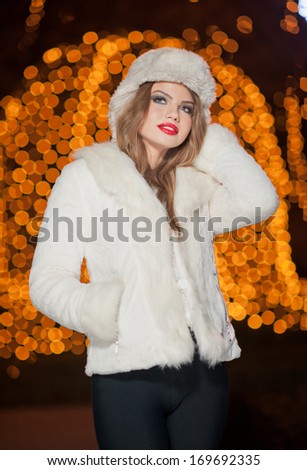 Fashionable lady wearing white fur cap and coat outdoor with bright Xmas lights in background. Portrait of young beautiful woman in winter style. Bright picture of beautiful blonde woman with make up