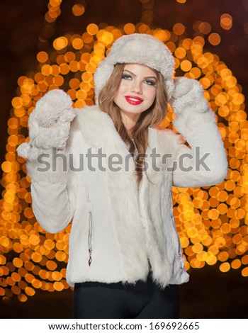 Fashionable lady wearing white fur accessories outdoor with bright Xmas lights in background. Portrait of young beautiful woman in winter style. Bright picture of beautiful blonde woman with make up