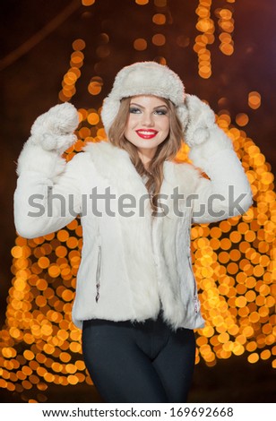 Fashionable lady wearing white fur accessories outdoor with bright Xmas lights in background. Portrait of young beautiful woman in winter style. Bright picture of beautiful blonde woman with make up