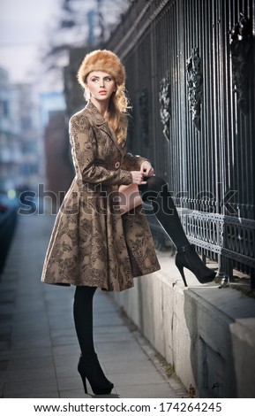 Attractive elegant blonde young woman wearing an outfit with Russian influence in urban fashion shot. Beautiful fashionable young girl with long legs and fur cap posing on street