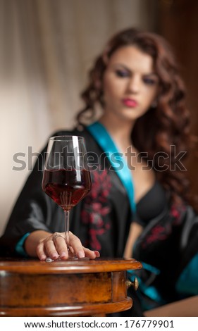 Beautiful sexy woman with glass of wine sitting on chair. Portrait of a woman with long curly hair posing challenging. Sexy brunette sitting near wood table with glass with red wine  in vintage scene