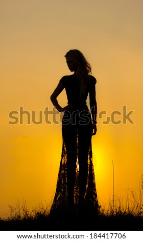 Silhouette of a young woman in long transparent dress in front of a beautiful sunset. Blonde in black lace dress posing outdoor in front of a beautiful sunset. Sensual girl with sun rising behind her.